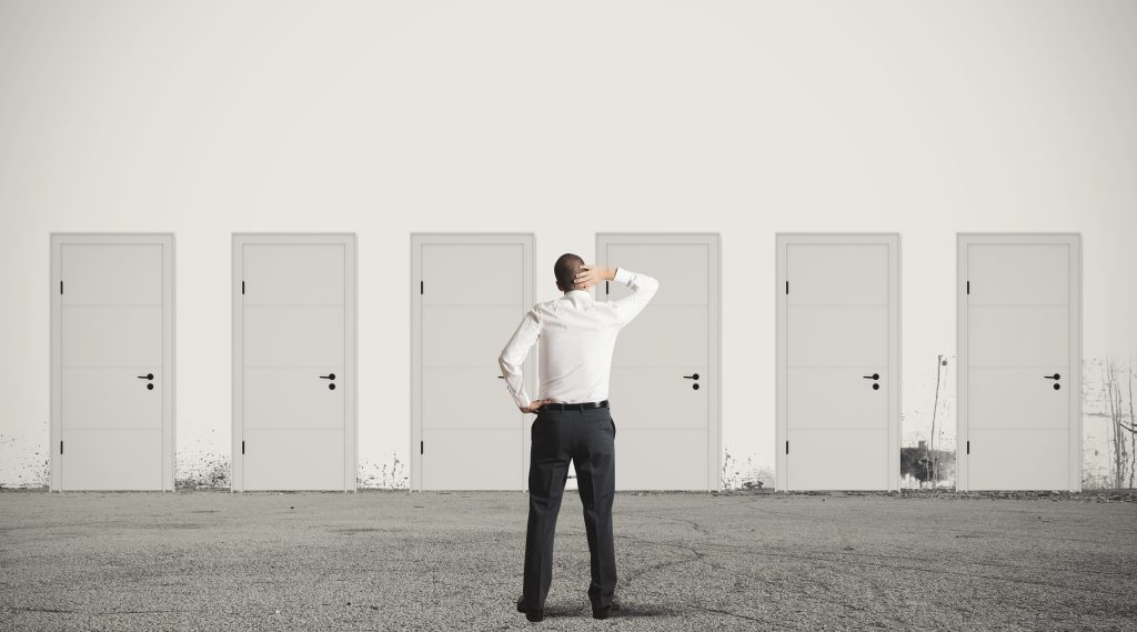 A person standing in front of 6 white doors seemingly struggling to make a decision