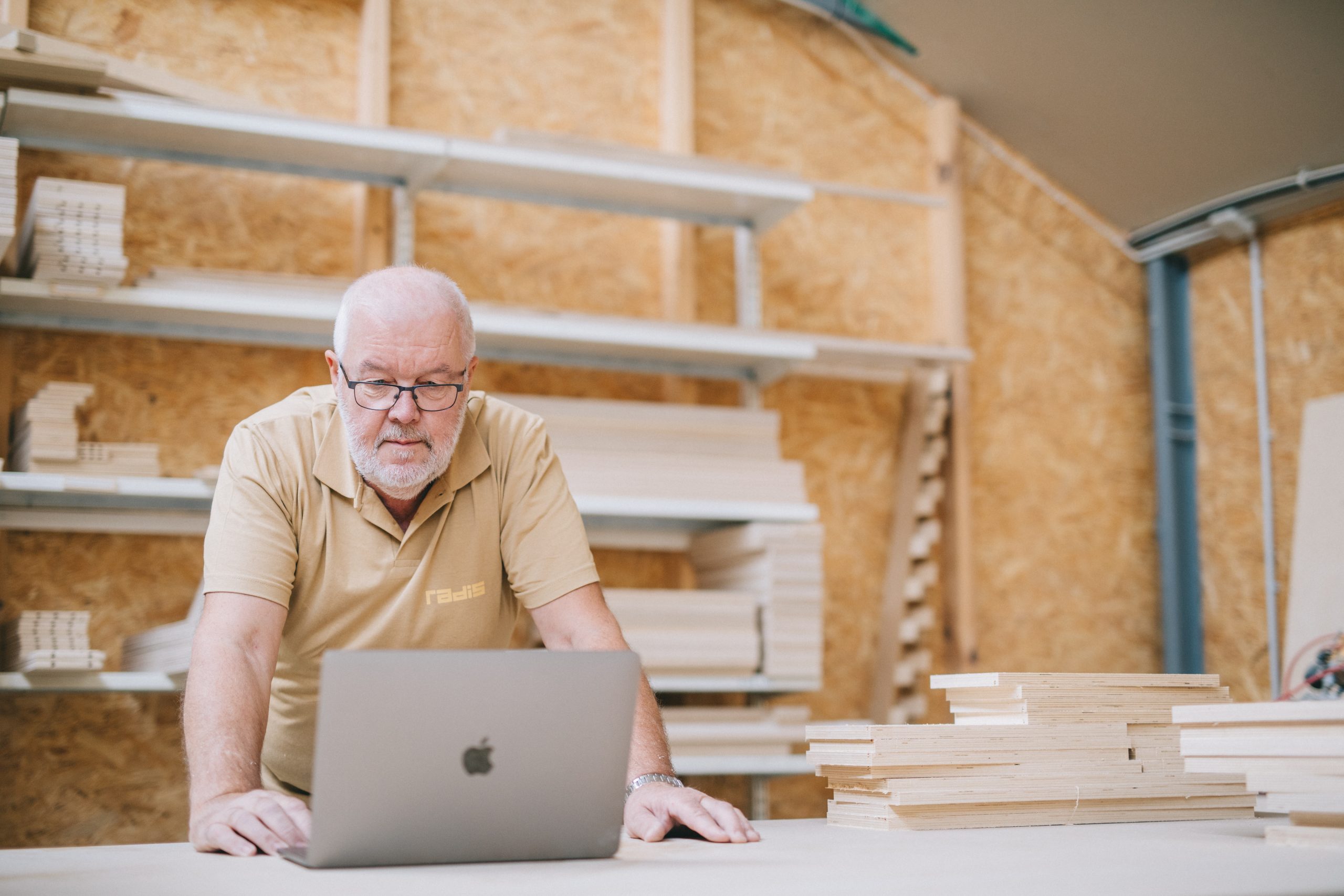 A worker in a beige polo shirt standing in a wood workshop looking at a laptop
