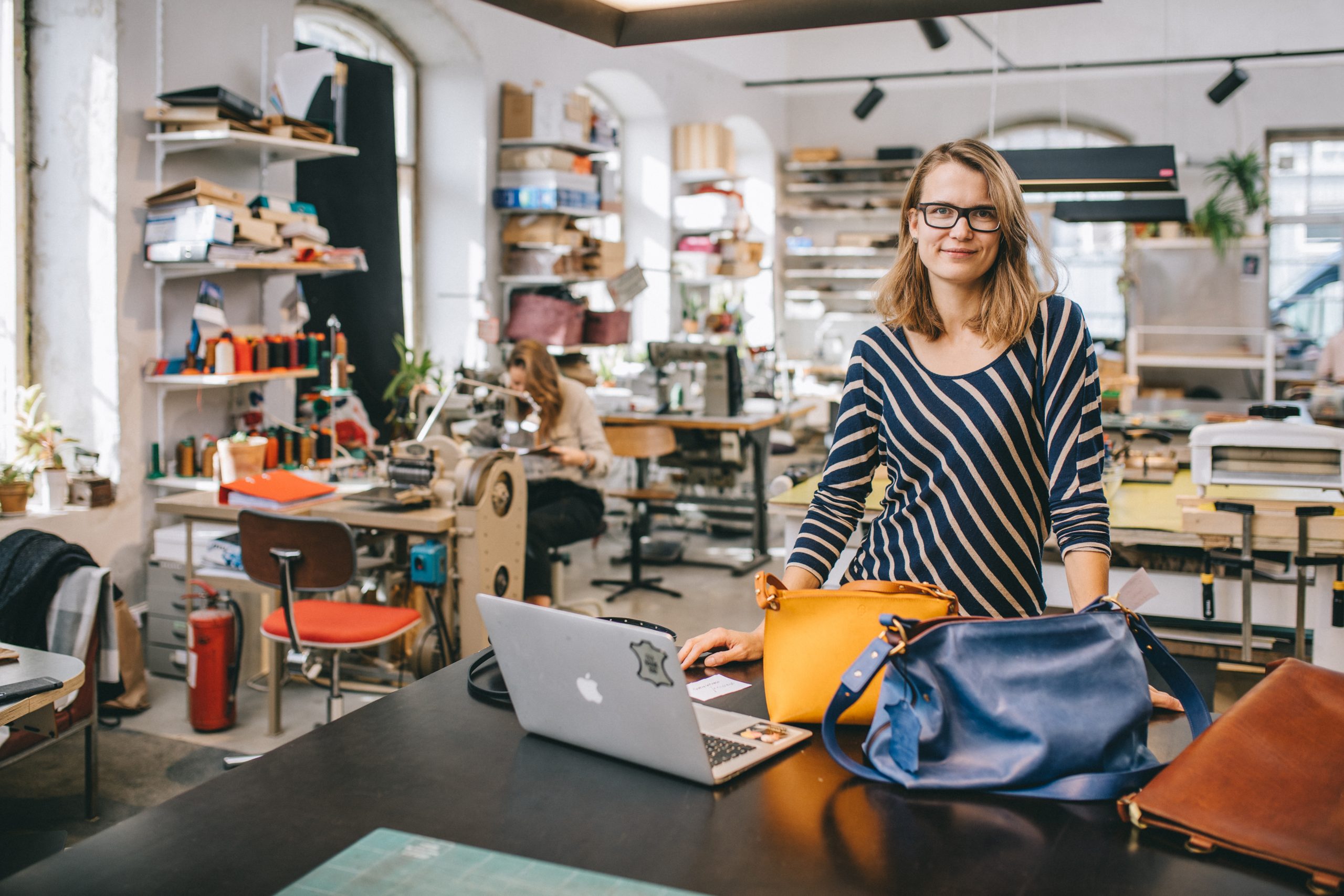 Leather maker stood at workstation with laptop and bags looking pleased they learned how to calculate selling costs. 