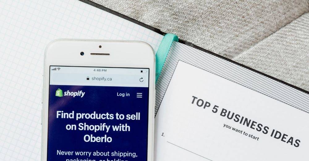 A planning diary with a pencil nearby sits on top of the table. A phone on the table shows the Shopify site as a part of the article on the best Shopify stores.