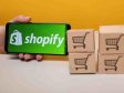 A handheld mobile device showing the Shopify logo next to small boxes with shopping cart symbols printed on