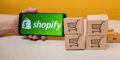 A handheld mobile device showing the Shopify logo next to small boxes with shopping cart symbols printed on