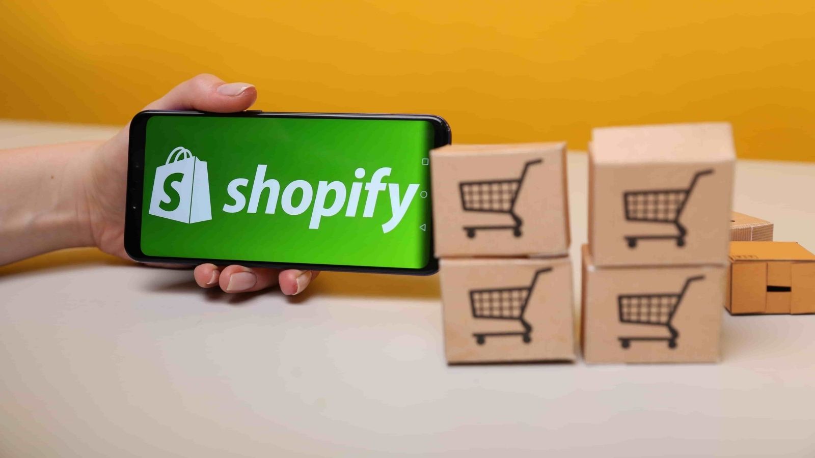 A handheld mobile device showing the Shopify logo next to small boxes with shopping cart symbols printed on