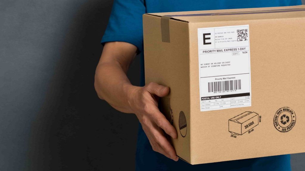 A person in a blue shirt holds a cardboard box with a Priority Mail Express label with 1D and 2D barcodes, indicating fast delivery service.