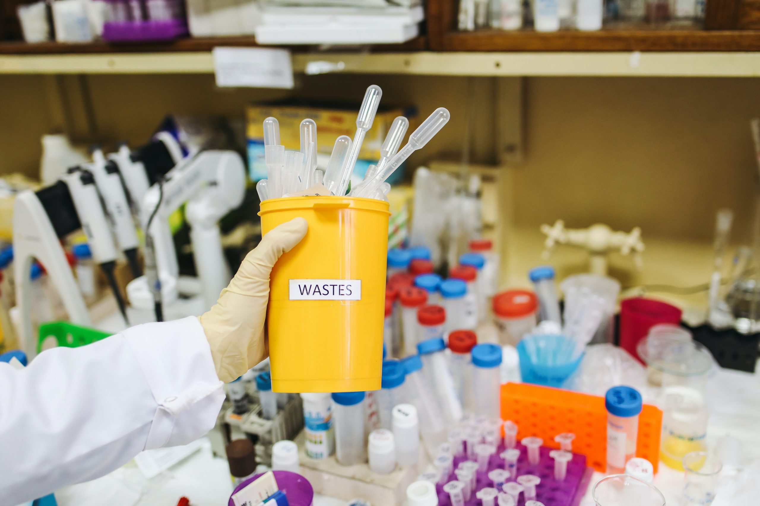 A worker in a laboratory holding a small waste bin with test tubes sticking out