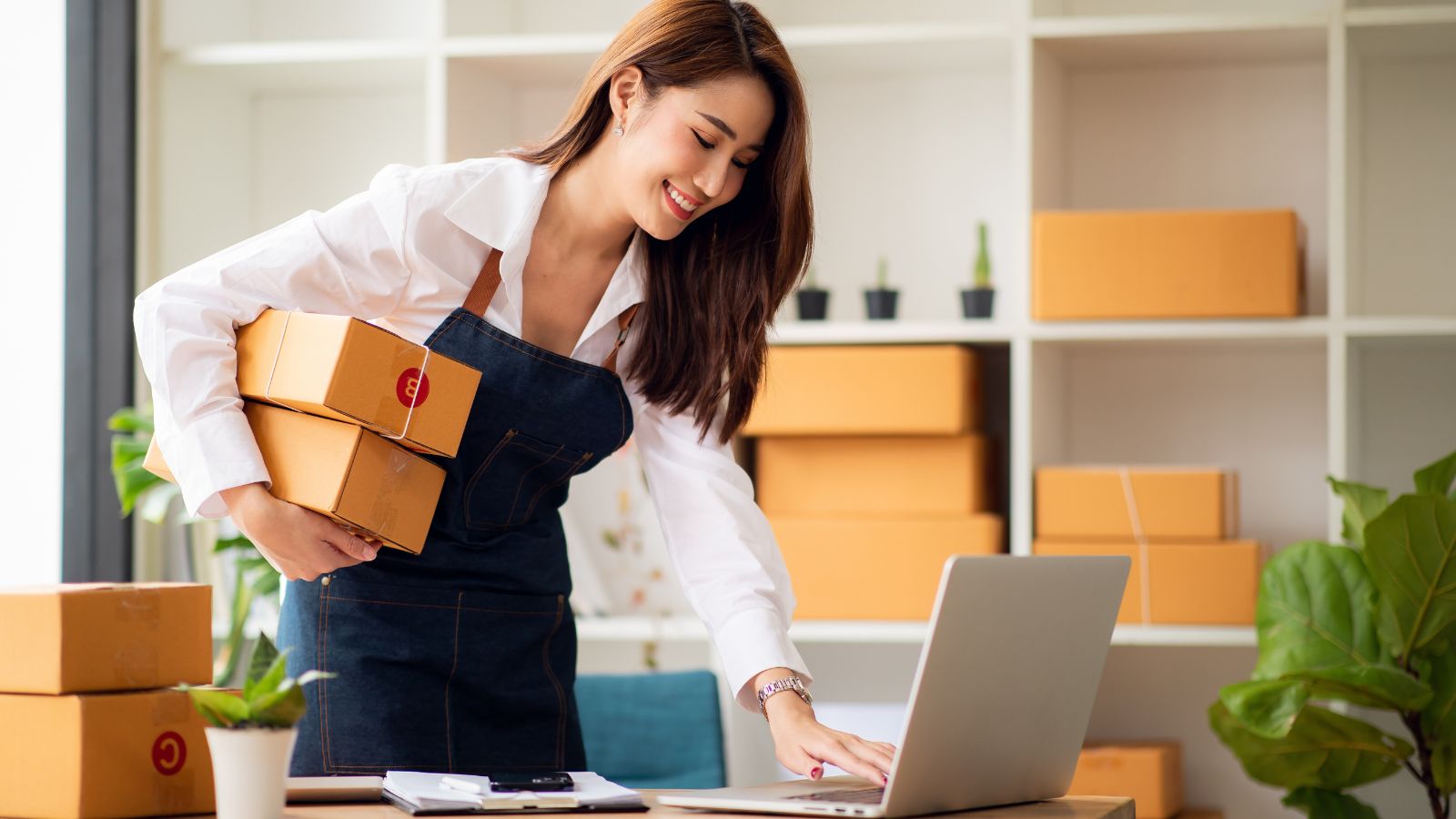 Manufacturer e-commerce in action, as a manufacturer handles packaged goods while updating the status of orders on her laptop.