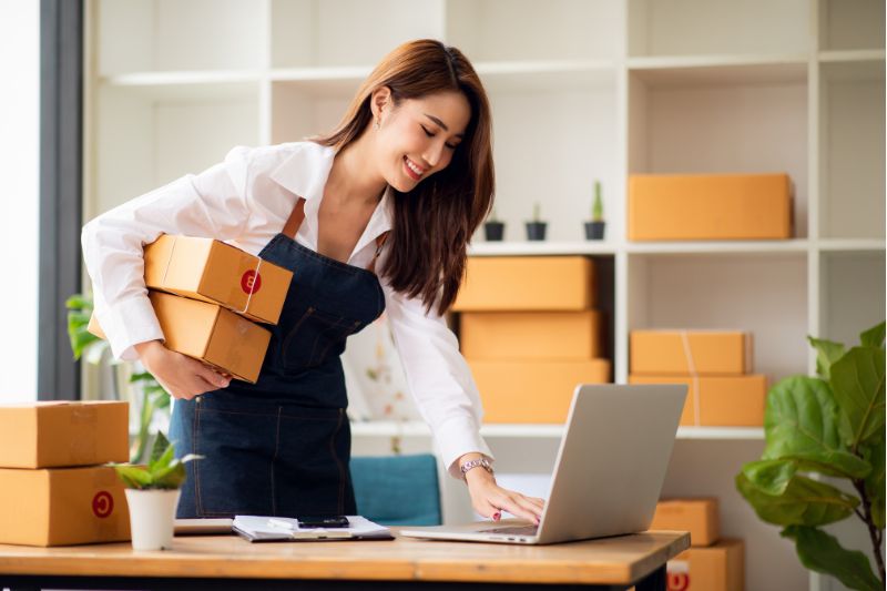 Manufacturer e-commerce in action, as a manufacturer handles packaged goods while updating the status of orders on her laptop.