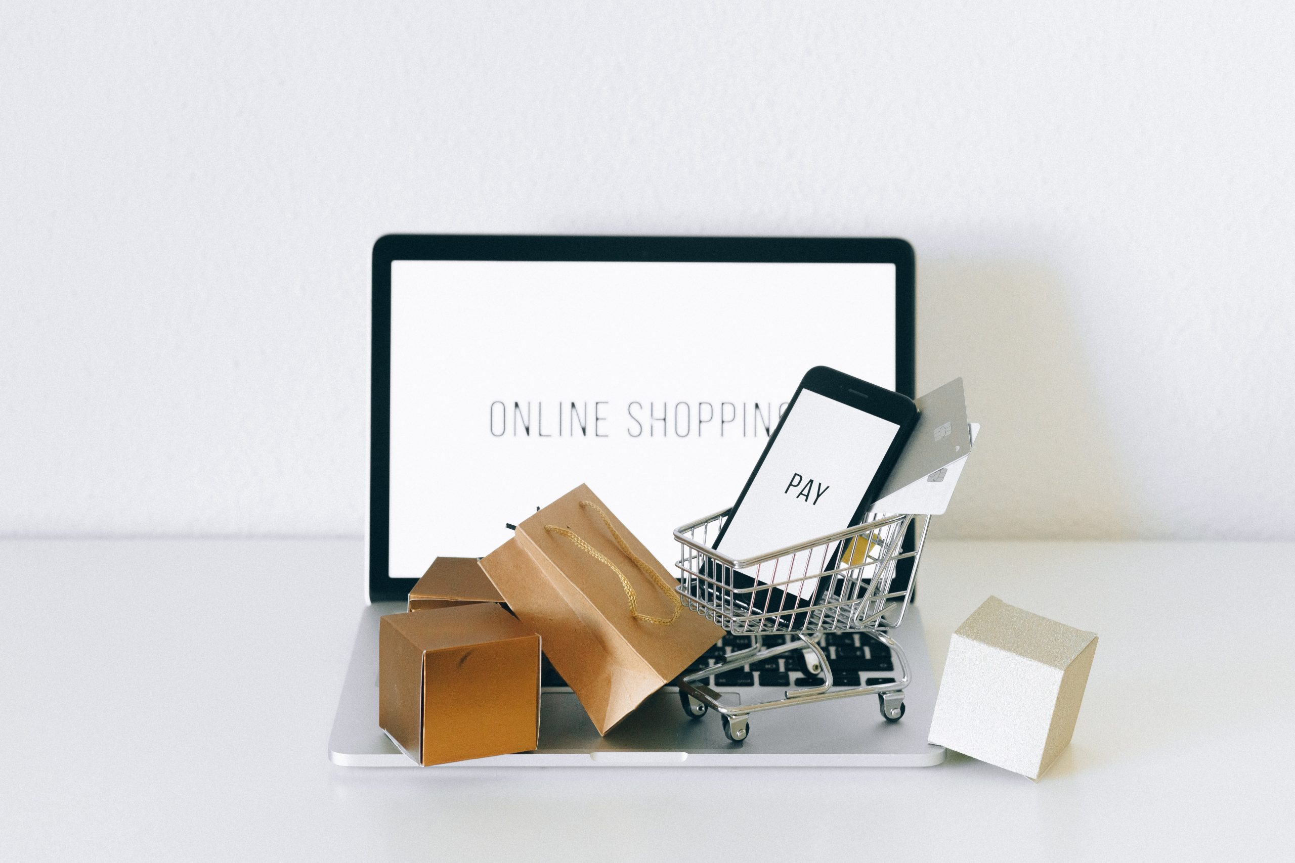 Laptop on a desk with words "online shopping" showing on the screen. Small shopping cart and boxes, bas laying on the keyboard.