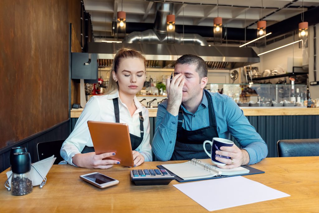 Two workers sitting at a table in an empty restaurant looking at a clipboard with worried faces