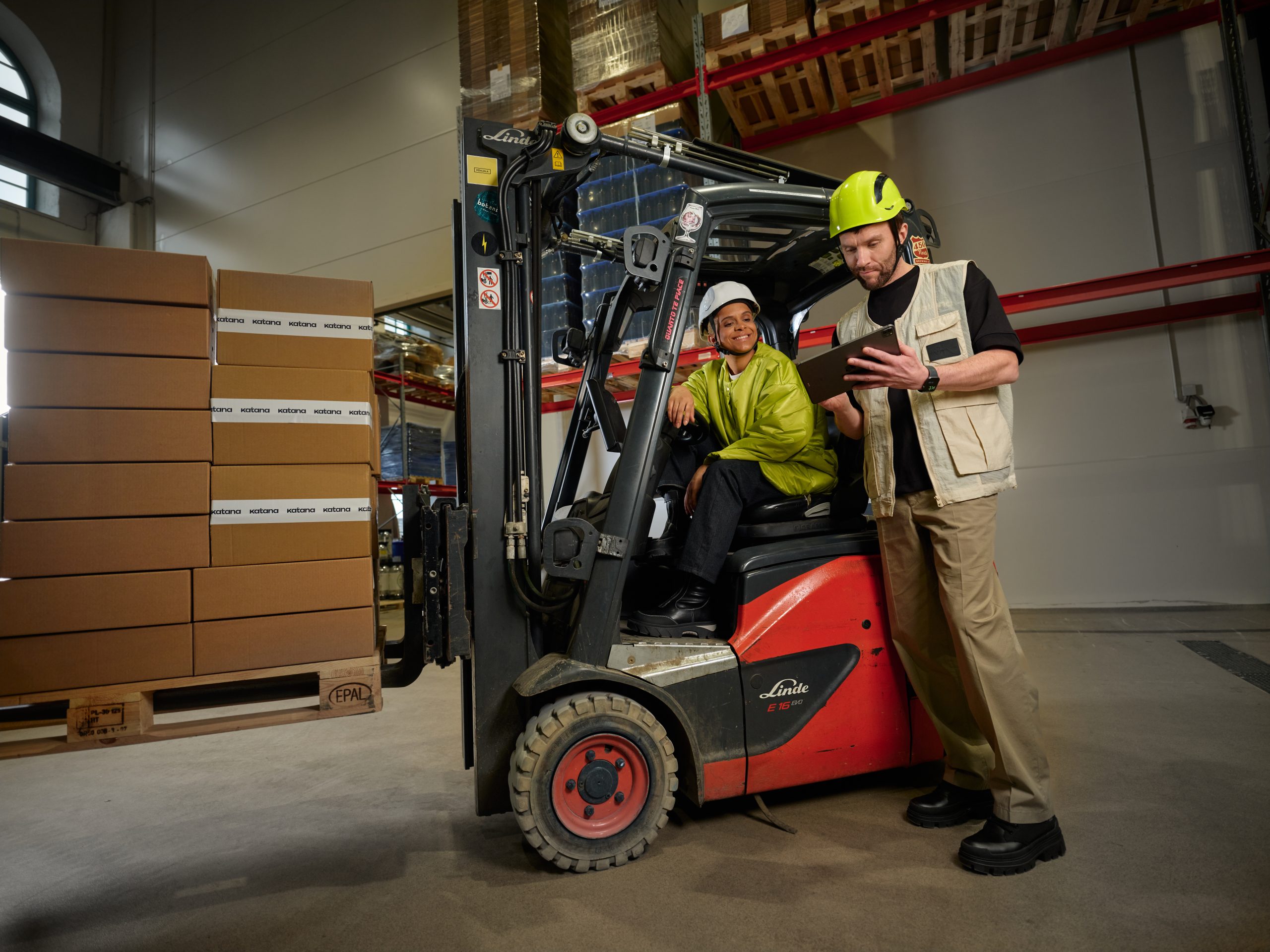 A worker sitting in a forklift talking to another worker while looking at their inventory management software on an iPad