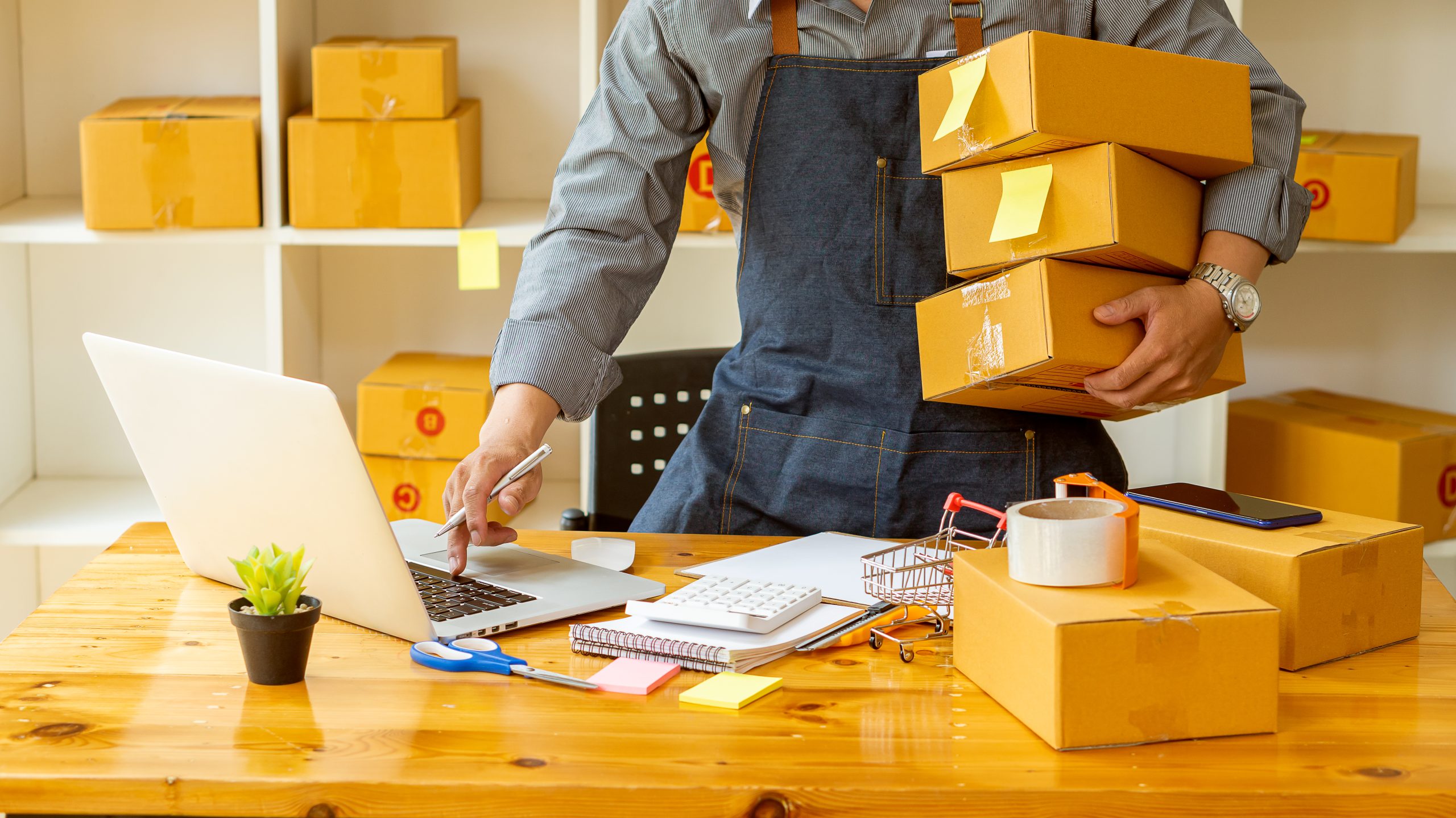 A man standing while holding multiple boxes and looking at a laptop