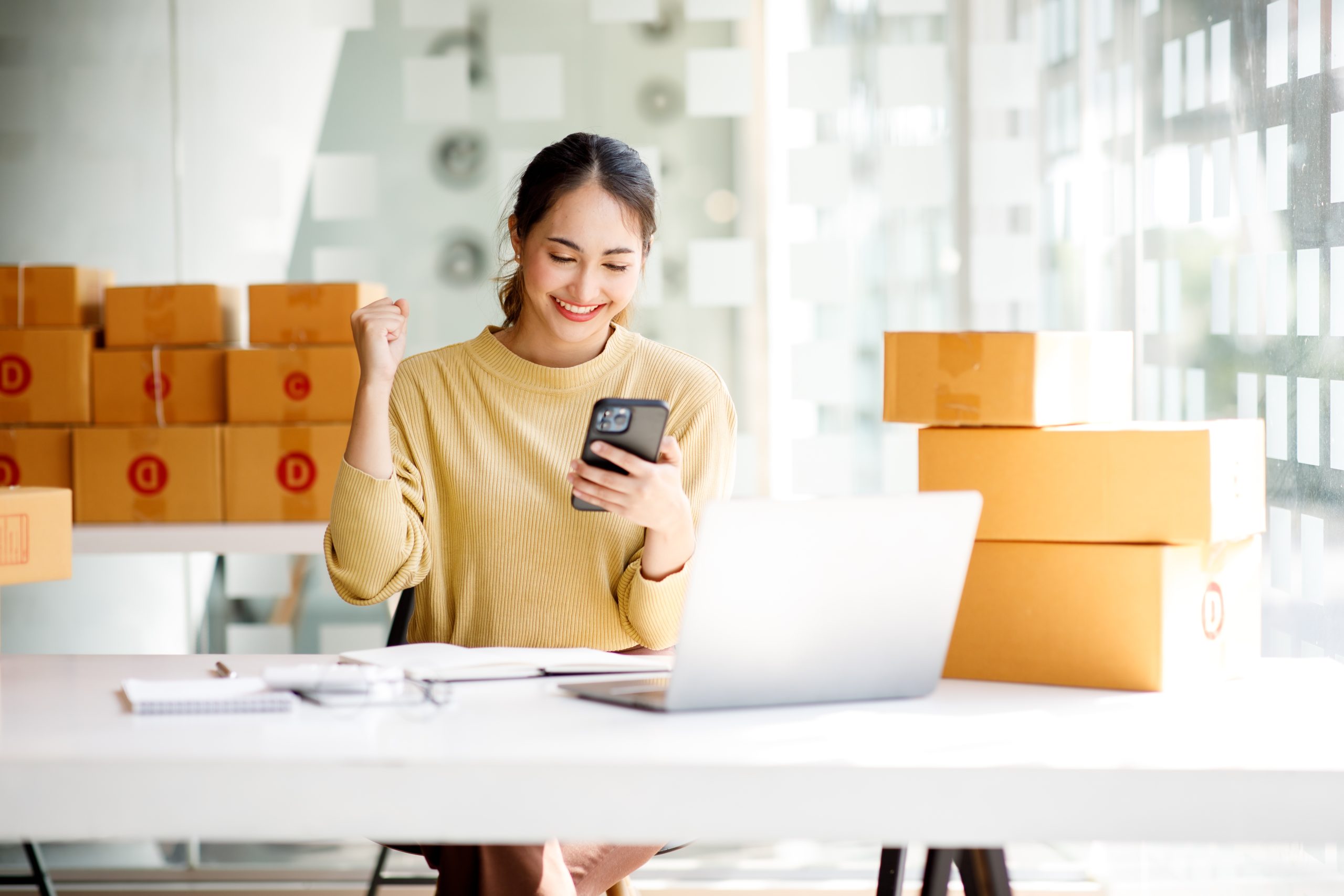 A woman sitting at a desk, looking at her phone with stacked boxes in the background