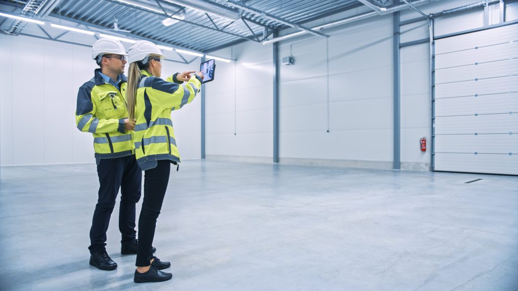 Two engineers standing in an empty warehouse and using an AR app on a tablet to figure out warehouse layout