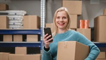 Warehouse manager holding smartphone and box in front of shelves, managing live inventory across locations